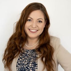 Sydney Comitz with long hair in a beige blazer and patterned top, against a white background.