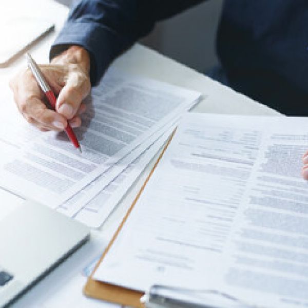 Man reviewing documents with a red pen near a laptop, highlighting sections for editing or approval.