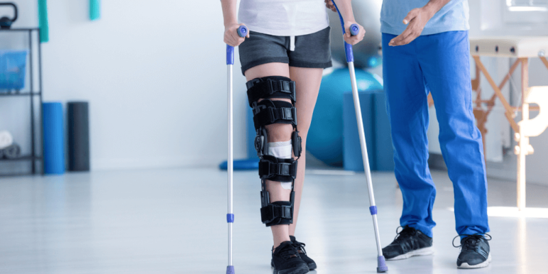 A person with a leg brace walks using crutches, assisted by another person, in a rehabilitation clinic with exercise equipment in the background.