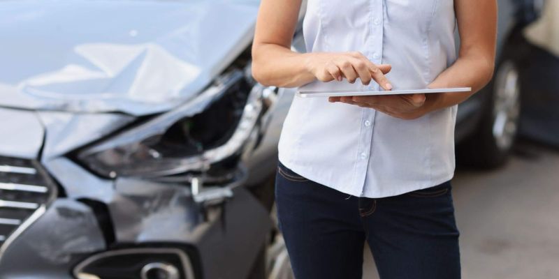 A woman is standing next to a car holding a laptop; the car seems to have been involved in a crash