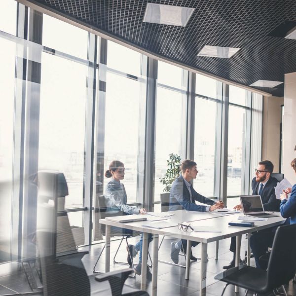 Business meeting in modern office with large windows, team discussing strategy around a table, laptops and documents visible.