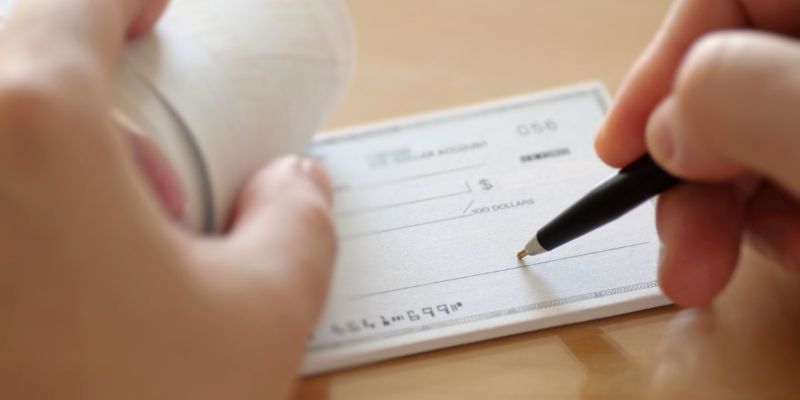A hand writing on a check using a pen. An adjacent hand holds a roll of receipts. The setting appears to be a table or desk.