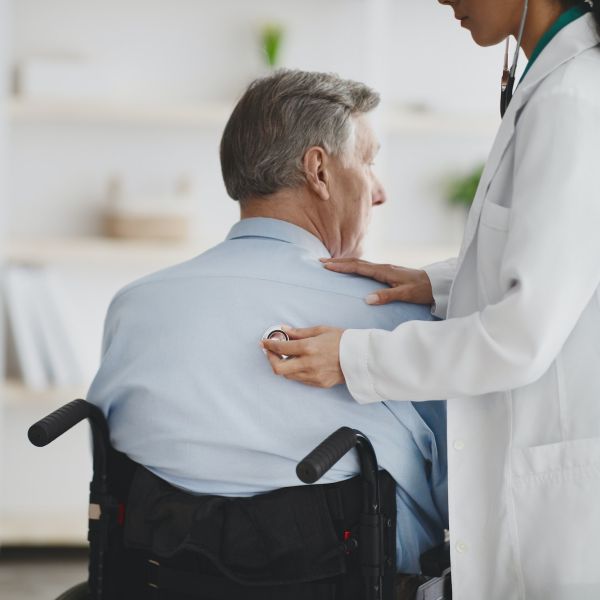 Female doctor using stethoscope on older man in wheelchair at home