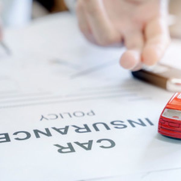 Red toy car on car insurance document with human hand holding a pen in background, symbolizing vehicle protection.