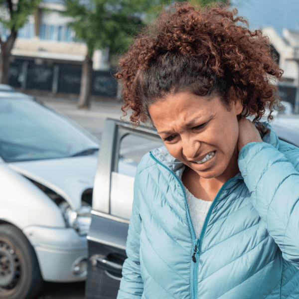 A woman grimaces, holding her neck, standing between two cars that appear to have collided in a parking lot surrounded by trees and buildings.