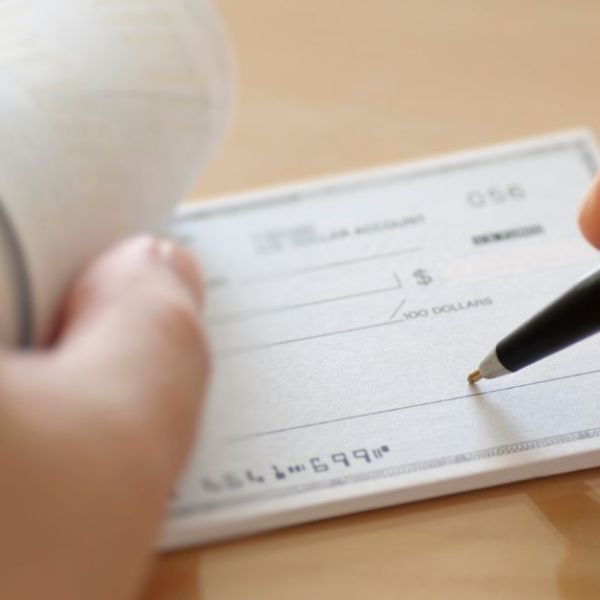 A hand writing on a check using a pen. An adjacent hand holds a roll of receipts. The setting appears to be a table or desk.