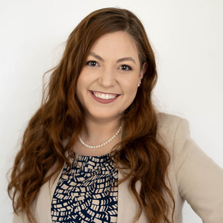 Sydney Comitz with long hair in a beige blazer and patterned top, against a white background.