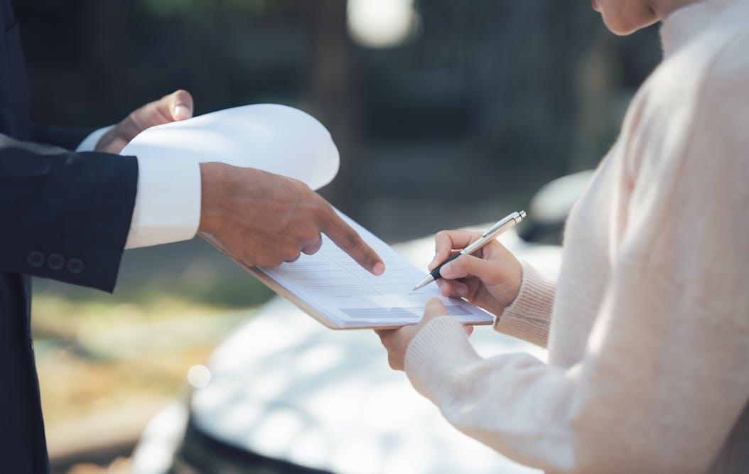 Hands hold a clipboard with paper; one person points, while another writes, outdoors near a white car, suggesting document review or signing.