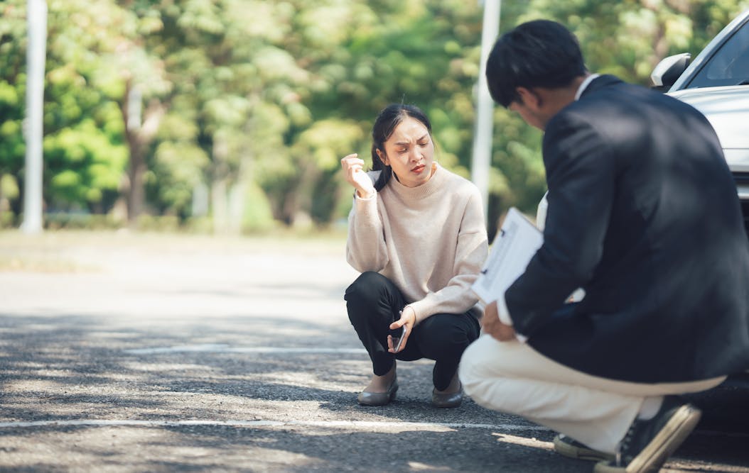 A woman crouches, holding a phone and gesturing, while a man kneels beside a car, holding papers. They're in an outdoor setting with blurred trees in the background.