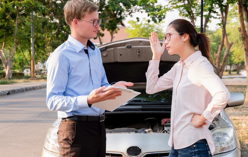 A person holding a clipboard talks to another person gesturing with their hand. They stand in front of a car with an open hood, surrounded by trees.