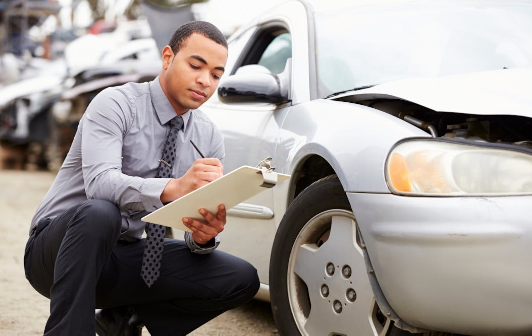 A person with a clipboard documents damage on a car with a smashed front bumper, in an outdoor setting. Text on the clipboard is not discernible.
