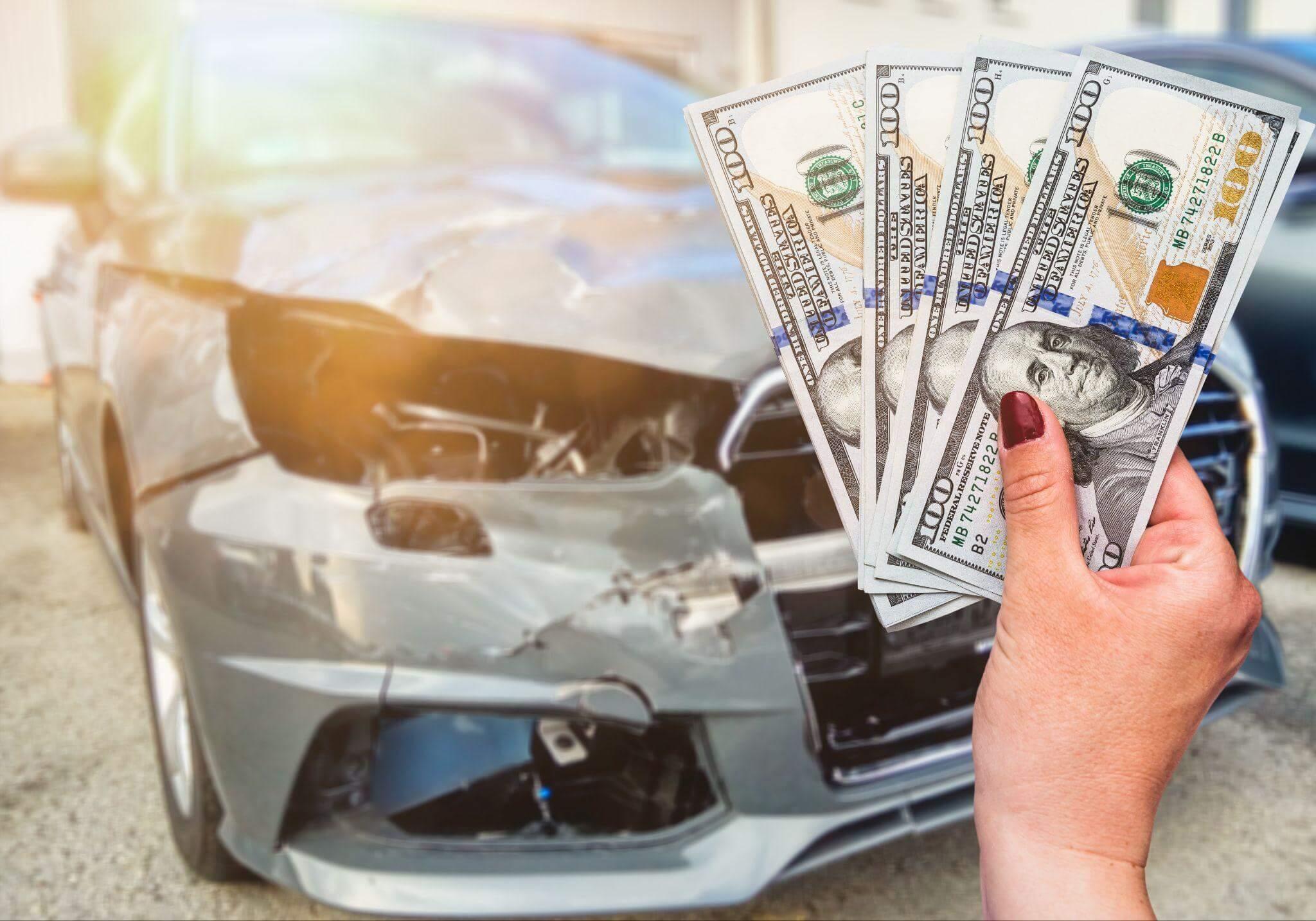 a person holding a bunch of money in front of a car, the car is very damaged indicating the money is the result of the accident